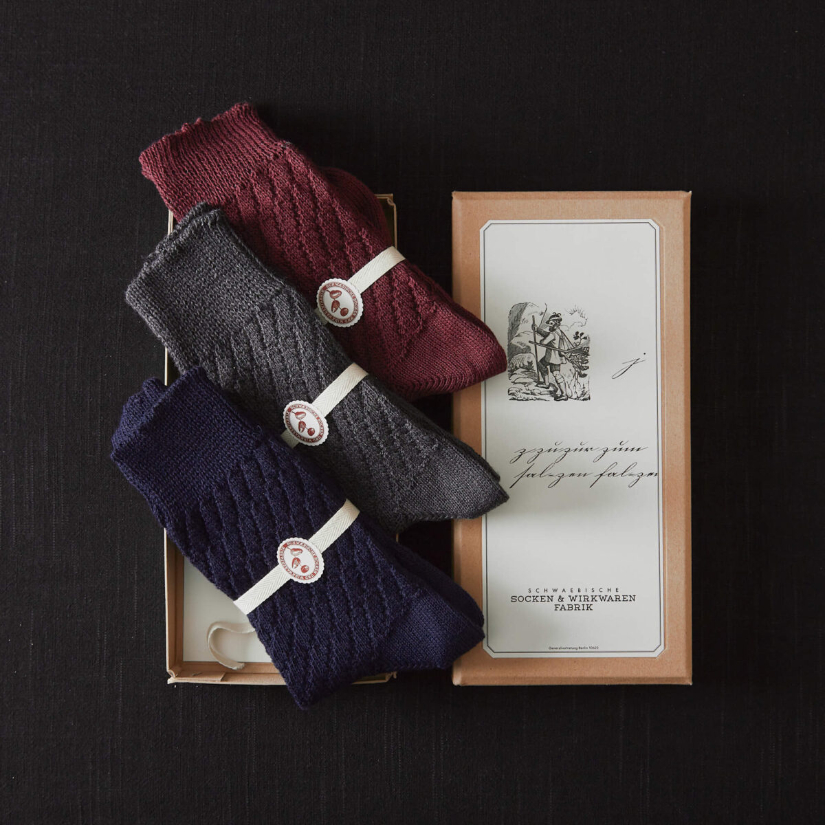 Zopfmuster-Socken im 3er Set von Frank Leder im Hausgemachtes Shop erhältlich in einer liebevoll gestalteten Papierverpackung mit handgeschriebenem Titel und feinen Linienzeichnungen. Die Socken sind in grau, navy und wein.
